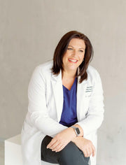 A smiling woman in a white coat with brown hair, showcasing a watch on her wrist, possibly related to a wellness or prenatal product.